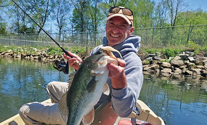 Middle-aged adult male wearing a tan cap and gray hooded sweatshirt holding a bass fish in one hand and the fishing reel in the other posing for a photo.