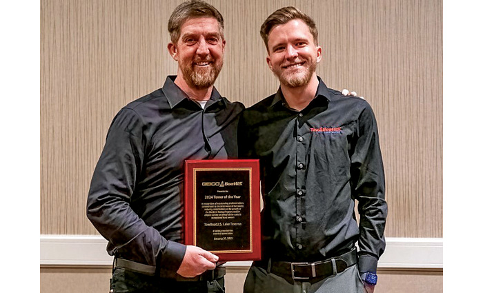 Two adult males wearing black long sleeve collared shirts posing for a photo while holding a plaque award.