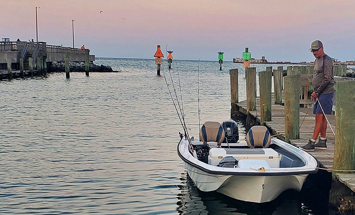 Adult male wearing a tan cap and long sleeve shirt with navy shorts tying a small white fishing boat to a wooden dock at dusk. 