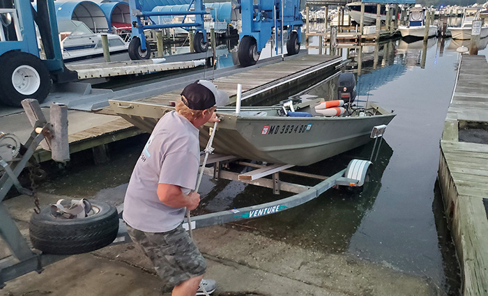 View from behind of an adult male wearing a gray shirt, black ballcap and shorts launching a silver fishing boat at a marina.