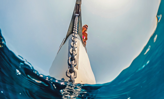View from below of a blonde female wearing sunglasses and a pink swimsuit looking down as an anchor on a chain enters clear blue waters.