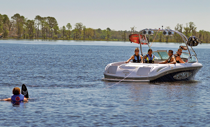 Four adults aboard a white boat on a lake with one about to water ski on a sunny day.