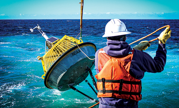 Back of an adult male wearing a blue hooded sweatshirt, white hard hat and orange life jacket pulling a large yellow buoy out of the ocean.