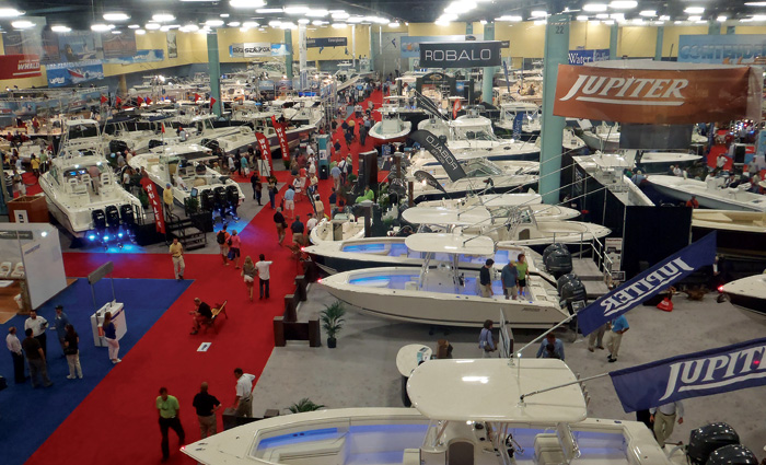 Aerial view of numerous boats and people walking on red carpet at an indoor boat show.