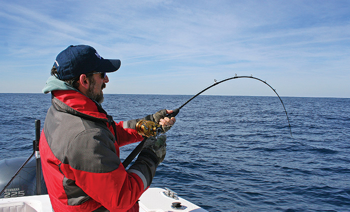 Adult man wearing a navy ballcap, sunglasses and red and gray jacket fishing off a white boat in open waters.