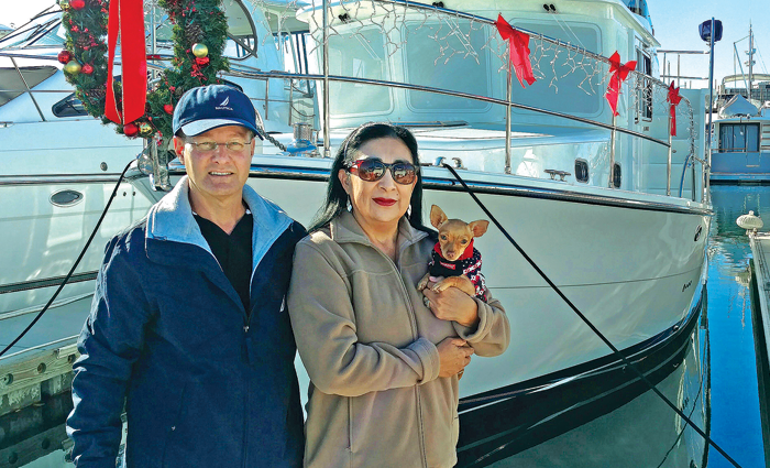 Adult male wearing a navy and blue jacket and navy ballcap next to an adult female wearing sunglasses and a tan jacket holding a small dog.